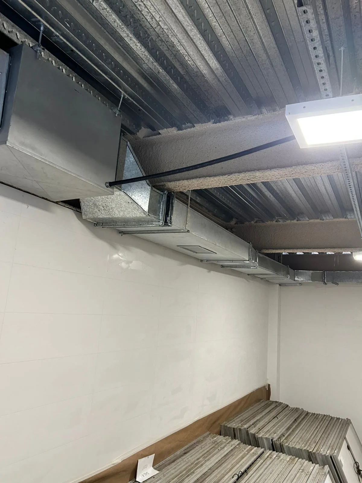 Duto retangular fixado ao longo de parede com revestimento cerâmico branco, com caixas de ventilação ao fundo
